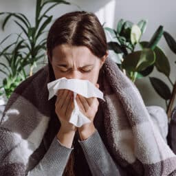 Woman wrapped in a blanket blowing her nose, affected by either seasonal allergies or the cold weather.
