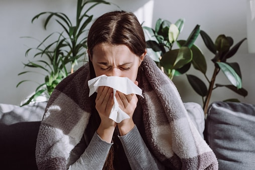 Woman wrapped in a blanket blowing her nose, affected by either seasonal allergies or the cold weather.