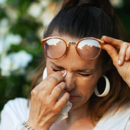 Woman struggles with watery eyes, a common summer allergy symptom. She wipes her eyes with a handkerchief.