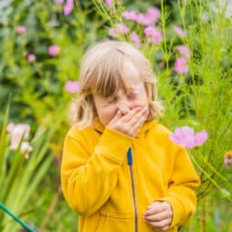 Little boy is sneezing outdoors in a flower garden wearing a yellow jacket.